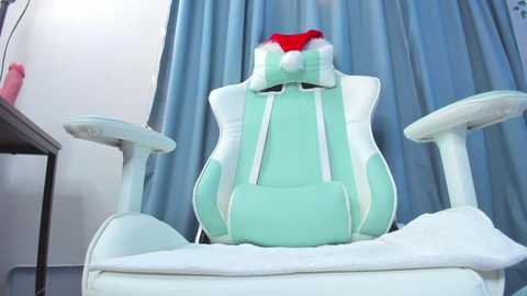 Media: Video of a light green gaming chair with a white headrest, featuring a red Santa hat on the headrest, against a blue curtain backdrop.