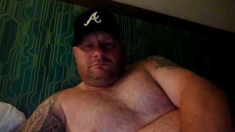 Media: A video of a shirtless, overweight, bearded man with glasses and a black Atlanta Braves baseball cap, sitting on a bed with green geometric-patterned wallpaper behind him.