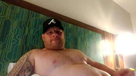 Video of a middle-aged, shirtless man with a large belly and tattooed arm, wearing a black cap with a white logo, sitting in a room with green patterned wallpaper and a lamp.