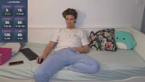 Media: A video of a young man with short brown hair, wearing a white t-shirt and light blue pajama pants, sitting on a light blue bed with a floral pillow and plush toy, holding a controller, in a room with a virtual reality headset screen.