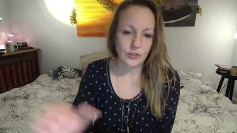 Video of a young woman with fair skin, long blonde hair, and a slim physique, wearing a black top with white polka dots. She's on a bed with a zebra-print duvet, in a room with a wooden headboard, a lamp, and a wall-mounted picture.