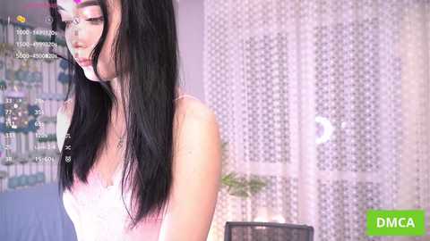 A video of a young woman with long black hair, wearing a pink lace camisole, standing in a room with a white patterned curtain.