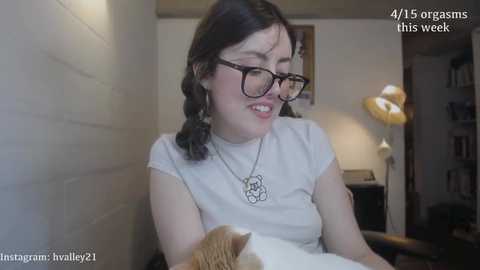 A young woman with fair skin, glasses, and short dark hair, wearing a white T-shirt, sits indoors, holding a cat. The room has a beige wall, a lamp, and a bookshelf.