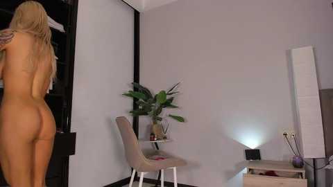 Video of a nude woman with long blonde hair standing in a modern, minimalist bedroom. She's facing away, revealing her bare buttocks. The room features a gray wall, a small potted plant, and a contemporary chair.