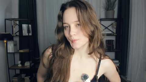 Video of a young woman with long brown hair, wearing a black bra, standing in a modern room with bookshelves and a potted plant.