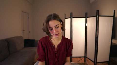 Video of a young woman with shoulder-length brown hair, wearing a maroon blouse, sitting indoors. The background features a gray sofa, a black lamp, and a wooden screen.
