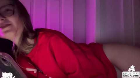 A video of a young woman with light skin and glasses, wearing a red sweater, lying on a bed with purple lighting.