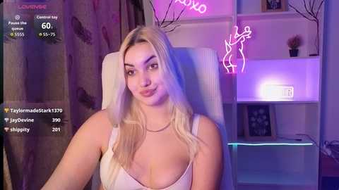 Video of a smiling blonde woman in a low-cut white top, seated in a white chair. Neon lights and a pink heart-shaped pillow in the background.