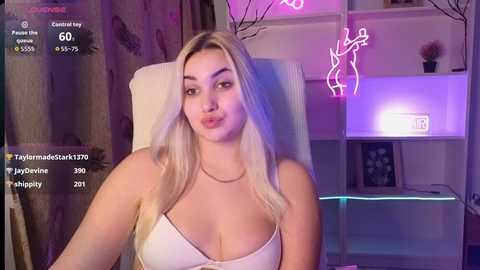 Video of a fair-skinned, blonde woman with long hair, wearing a white bra, seated in a dimly lit room with purple neon lights and a clock.