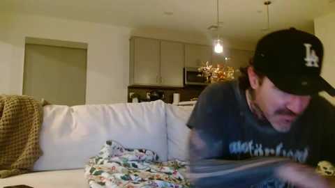 A video of a man wearing a black hat and T-shirt, lying on a white couch in a cozy living room with a TV, kitchen, and wine glasses in the background.