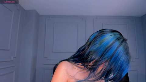Video of a person with long, vibrant blue hair, partially obscuring their face, in a room with grey walls and closed doors.