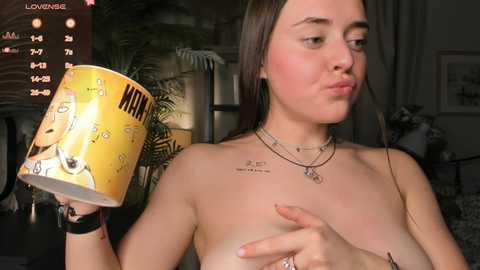 Video of a young, light-skinned woman with long brown hair, topless, wearing a yellow mug with cartoon characters, kissing her left breast, in a dimly lit room.