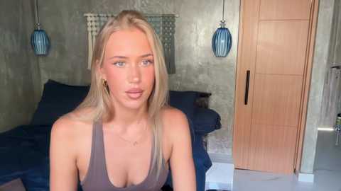 Video of a blonde woman with fair skin, wearing a grey tank top, sitting on a bed in a modern, minimalist bedroom with blue bedding, wooden door, and pendant lights.