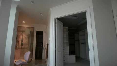 Video of a modern, empty, unfinished bathroom with beige walls, glass shower, white chair, and open door revealing a closet with boxes.