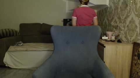 Video of a woman in a red top, standing with her back to the camera, in a living room with a blue armchair, beige sofa, patterned wallpaper, and wooden furniture.