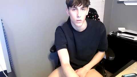 Video of a young man with fair skin, dark hair, wearing a black t-shirt, sitting in a black office chair against a grey wall, with a desk and window partially visible.