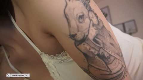 Video of a person's upper arm with a detailed, black-and-grey tattoo of a dog's face, wearing a white lace-trimmed tank top. Background shows a bedroom with white walls and framed pictures.