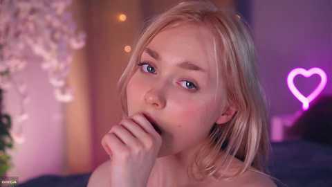 Video of a young, blonde woman with fair skin and blue eyes, holding her finger to her lips in a seductive pose. Background features pink heart-shaped neon lights and blurred cherry blossoms.