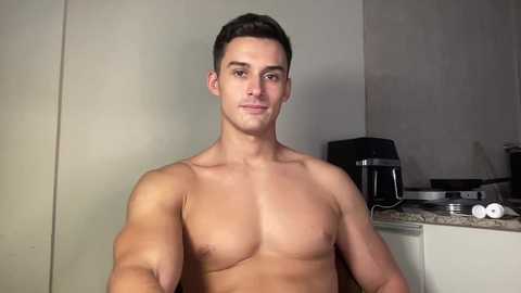 Video of a shirtless, muscular man with short brown hair, standing in a kitchen with granite countertops, a black coffee maker, and white appliances.