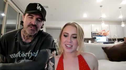 Video of a tattooed man with a mustache, wearing a black LA Dodgers cap, and a woman with blonde hair in a red halter top, sitting on a couch in a modern kitchen.