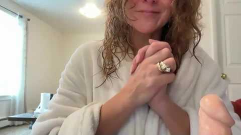 Video of a smiling, fair-skinned woman with wet, curly brown hair, wearing a white bathrobe and holding her hands clasped near her chest, standing in a brightly lit bathroom with a window and door visible in the background.