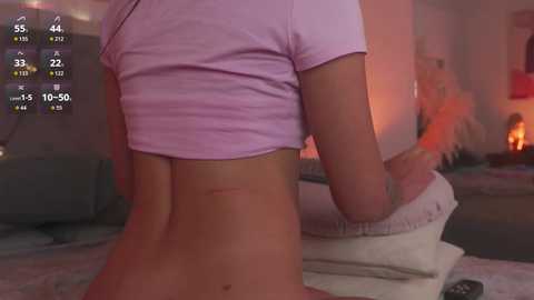 A video of a fair-skinned woman in a cropped pink t-shirt, kneeling on a bed, with a fireplace and cozy room in the background.