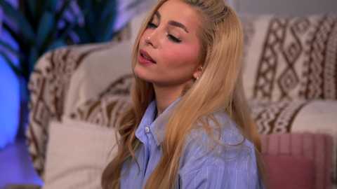 Video of a blonde woman with long hair, closed eyes, wearing a blue button-down shirt, standing against a beige couch with patterned pillows.