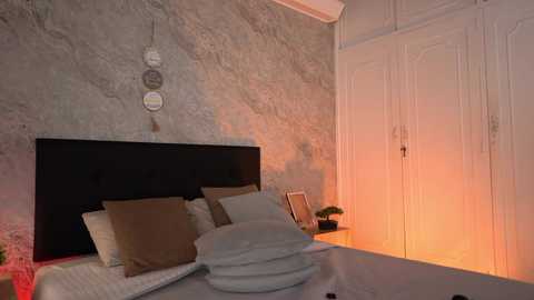 Video of a cozy bedroom with a black tufted headboard, beige pillows, white sheets, a dreamcatcher wall decoration, and a warm glow from a nearby closet.