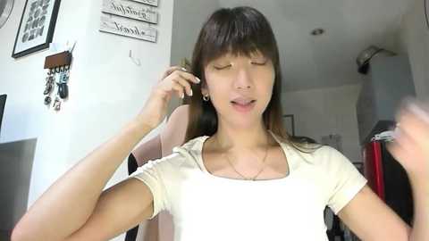 Video of a young Asian woman with straight brown hair, wearing a white top, holding a phone to her ear, in a modern living room with white walls and framed art.