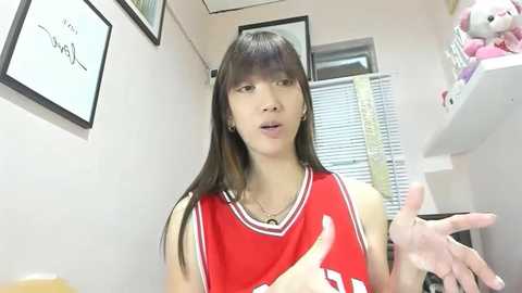 Video of an Asian woman with long black hair in a red basketball jersey, talking animatedly in a bright, tidy room with white walls, framed artwork, and a pink stuffed bear.