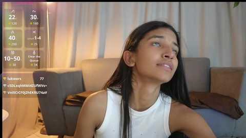 Media: Video of a young woman with long black hair, light skin, wearing a white tank top, sitting on a couch in a cozy living room.