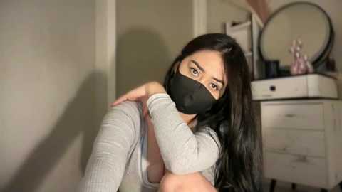 Video of an East Asian woman with long black hair, wearing a black face mask and gray sweater, seated on the floor in a softly lit room with a vanity and mirror in the background.