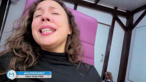 Video of a Caucasian woman with curly hair, wearing a black top, smiling with her mouth open, sitting on a pink cushioned chair in a wooden-framed room.
