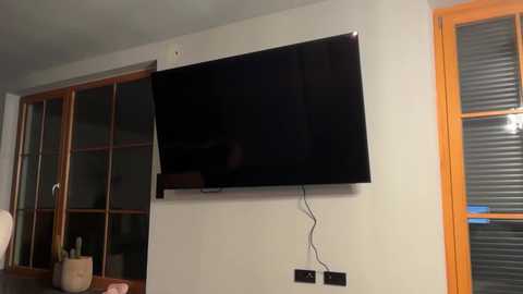 Video of a modern living room featuring a large flat-screen TV mounted on a beige wall, with wooden-framed windows and a white chair visible in the background.