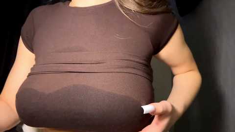 Video of a pregnant woman with fair skin, wearing a tight, dark brown t-shirt that accentuates her large, round belly. Her hands rest on her abdomen. The background is indistinct and dark.