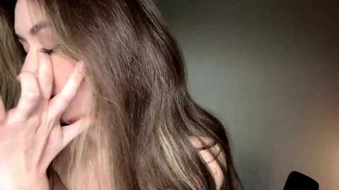 Video of a young woman with long, wavy brown hair partially covering her face, gently touching her lips with her right hand. The background is blurred with muted green and brown tones.