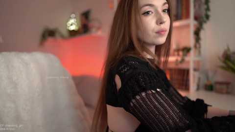 A video of a fair-skinned woman with long brown hair, wearing a black crochet dress, sitting in a cozy, softly lit room with bookshelves and plants.
