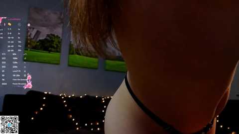 Video of a person's back in dimly lit room, wearing black underwear, with a calendar and a blurred image in the background.