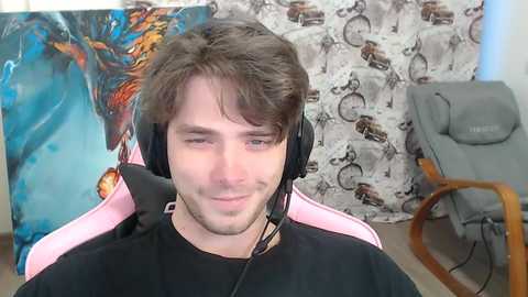 Video of a young man with brown hair, wearing a black shirt, headphones, and a pink gaming chair, in a room with a blue dragon painting and a gray recliner.