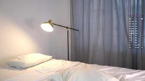 A minimalist bedroom with a white bed, a single white pillow, and a modern black floor lamp with a large, bright white bulb. Gray curtains cover the window, partially revealing a cityscape.