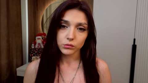 Video of a young woman with long dark hair, fair skin, and dark eyes, wearing a pink top, in a bedroom with wooden walls, a red plush toy, and a mirror.