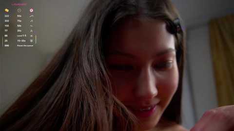 Video of a young Asian woman with long, dark hair, smiling, in a dimly lit room. Background shows a yellow curtain and a clock displaying 2:27 PM.