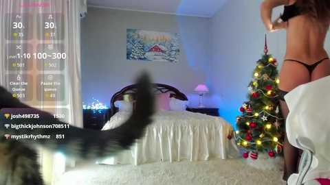 Video of a woman in black lingerie, holding a lit Christmas tree, in a bedroom with a white bed, a framed painting, and a digital display showing chat messages.