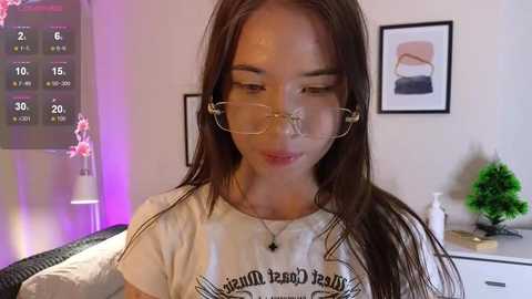 Video of a young woman with long dark hair and glasses, wearing a white t-shirt, in a cozy, dimly-lit room with pink lighting, a calendar, and art on the wall.