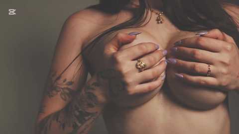 Video of a topless woman with long dark hair, covering her medium-sized breasts with her hands, adorned with purple nail polish and tattoos on her arms.