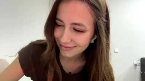 A video of a young woman with long, straight brown hair, wearing a black t-shirt, smiling gently, and standing in a white-tiled bathroom.