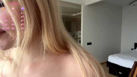 Video of a blonde woman with long hair, seen from the side, in a modern bedroom with white walls, bed, and open closet.