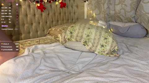 Video of a cozy bedroom with a plush beige tufted headboard, a white quilted bedspread, and decorative pillows. Red Christmas ornaments hang above the headboard.