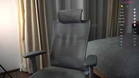 Media: Video of a modern office chair with a gray mesh backrest and a gray padded seat, placed in a minimalist bedroom with beige curtains and a gray bedspread.