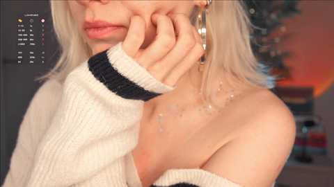 Video of a fair-skinned woman with platinum blonde hair, wearing a white off-shoulder sweater, large gold hoop earrings, and a delicate necklace. Her left hand rests on her cheek, and the blurred background suggests a cozy indoor setting.